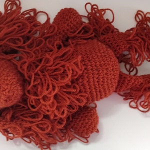 May include: A crocheted stuffed animal in the shape of a lion with a red-brown color. The lion has a long mane of yarn.