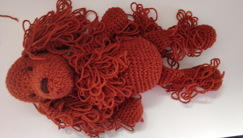 May include: A crocheted stuffed animal in the shape of a lion with a red-brown color. The lion has a long mane of yarn.