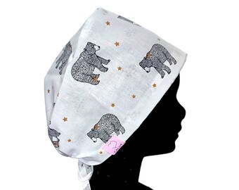 May include: White surgical cap with a repeating pattern of gray bears and gold stars. The cap is tied at the back with white ties. A pink tag with the text "CALOT" is attached to the cap.