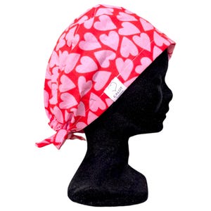 Peut inclure: Un bonnet de chirurgien rouge et rose à motifs de cœurs. Le bonnet est noué dans le dos et comporte une étiquette blanche avec le texte "Calot Creation". Le bonnet est présenté sur une tête de mannequin noire.