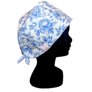 Peut inclure: Un bonnet de chirurgien à motif floral bleu et blanc. Le bonnet présente un motif répété de grandes fleurs et de feuilles bleues sur fond blanc. Une petite étiquette rose avec le texte "CALOT" est visible. Le bonnet est noué à l'arrière.