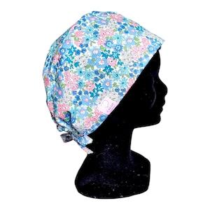 Può includere: Un berretto chirurgico bianco con un motivo floreale blu e rosa. Il berretto ha un laccio sul retro e una piccola etichetta rosa con il testo "CALOT" su di esso.