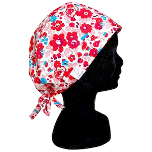 Op de afbeelding: Een scrubmuts met bloemenpatroon in rood, roze en blauw op een witte achtergrond. De muts is aan de achterkant vastgebonden met bijpassende stoffen bandjes. De tekst "CALOT" is zichtbaar op een klein wit label. De muts wordt getoond op een zwarte mannequin hoofd.