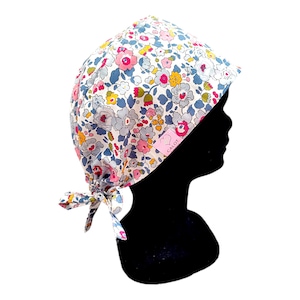 May include: A white surgical cap with a floral print in shades of pink, blue, and yellow. The cap has a tie at the back and a small pink label with the text "CALOT" on the side.