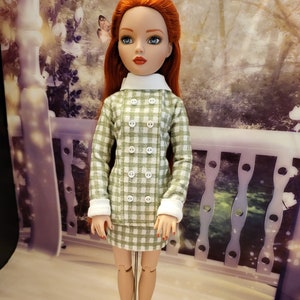 Könnte beinhalten: Eine Puppe, die ein grün-weiß kariertes Kleid mit weißen Knöpfen und einem weißen Kragen trägt. Die Puppe hat rote Haare und blaue Augen.