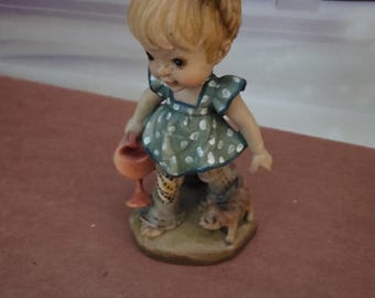 Puede incluir: Figura de cerámica de una niña con cabello rubio y un gran lazo. Lleva un vestido verde con lunares y leggings estampados, sosteniendo una pequeña copa. Un perro pequeño está a su lado. La figura está sobre una base marrón.