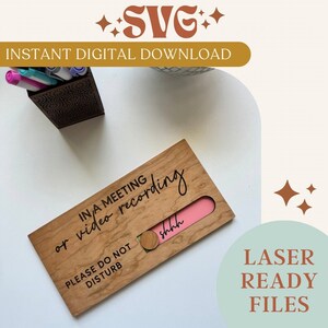 Op de afbeelding: Een houten bord met een schuifpaneel waarop staat "In een vergadering of tijdens video-opnames, niet storen". Het paneel schuift om een roze gedeelte te onthullen met de tekst "ssst".