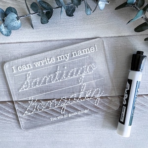 May include: A clear plastic dry-erase board with the text "I can write my name!" and the names "Santiago" and "Hong" written in dotted lines. A white dry-erase marker with black accents is next to the board. The text "You will do great things!" is printed below the names.