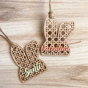 May include: Two wooden bunny-shaped ornaments with intricate cut-out designs. The ornament on the left has the name "Santi" written in light yellow paint. The ornament on the right has the name "Valentina" written in light pink paint.