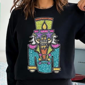 May include: Black sweatshirt featuring a colourful glitter-covered nutcracker design. The nutcracker is wearing a blue jacket with gold buttons and a red sash. The design is outlined with white stitching.