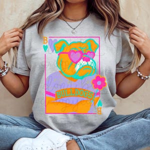 Peut inclure: T-shirt gris avec un bouledogue de dessin animé portant des lunettes de soleil en forme de cœur. Le motif comprend le mot "BULLDOGS" dans une bannière, une fleur rose et une palette de couleurs violet et orange. Le t-shirt a des manches courtes.