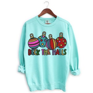 May include: A turquoise blue sweatshirt with a Christmas design featuring five colorful ornaments and the text "Deck the Halls".