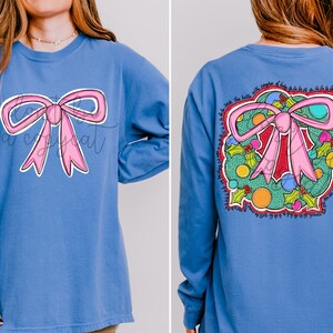 May include: A blue long-sleeve shirt featuring a pink bow graphic on the front and back. The back design includes a wreath with holly, colorful ornaments, and text. The shirt is a comfortable, casual style.