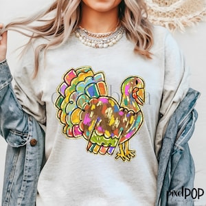 May include: A white long-sleeved shirt with a colorful cartoon turkey design. The turkey is outlined in gold and has a brown body with colorful feathers.