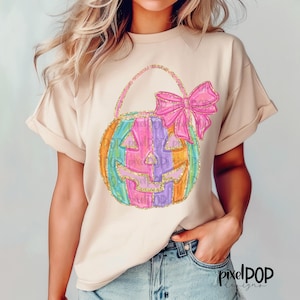 Puede incluir: Una camiseta beige claro con un diseño de calabaza colorida. La calabaza está rayada con rosa, morado, verde, naranja y amarillo. Tiene un lazo rosa en la parte superior y detalles de purpurina dorada.