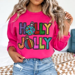 Peut inclure: Un sweat-shirt rose avec les mots "HOLLY JOLLY" en lettres scintillantes colorées avec un contour cousu. Chaque lettre a un motif d'ornement de Noël.