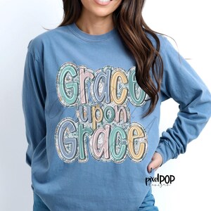 May include: A light blue long-sleeved shirt with a floral design and the text "Grace upon Grace" in a colorful, whimsical font.