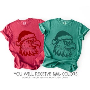 May include: Two comfort color t-shirts, one crimson and one light green, each featuring a graphic of an eagle's head wearing a Santa hat. The text "YOU WILL RECEIVE both COLORS" is displayed below the shirts.