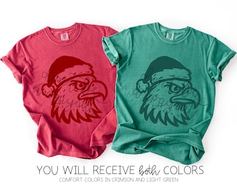 May include: Two comfort color t-shirts, one crimson and one light green, each featuring a graphic of an eagle's head wearing a Santa hat. The text "YOU WILL RECEIVE both COLORS" is displayed below the shirts.