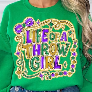 Może przedstawiać: Zielona bluza z nadrukiem "LIFE OF A THROW GIRL" w kolorze fioletowym, złotym i zielonym. Projekt zawiera kokardę, gwiazdki i obramowanie z koralików. Bluza jest w jednolitym zielonym kolorze.