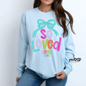May include: A light blue long-sleeved shirt with a colorful graphic design that says "So Loved" with a bow and the text "John 3:16".
