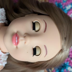 以下が含まれることがあります： 長いブロンドの髪、閉じられた目、そして描かれた眉毛を持つ人形。人形は白いレースのドレスを着ています。