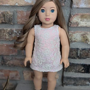 以下が含まれることがあります： ピンクと白のレースのドレスとゴールドのサンダルを履いた人形。人形は長い茶色の髪と青い目をしています。