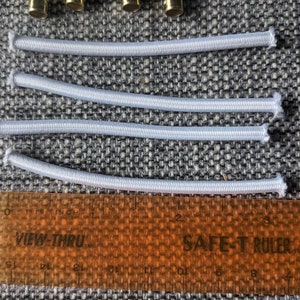 May include: Four white elastic bands laid out on a gray fabric surface with a ruler underneath. The ruler is orange and has the words "VIEW-THRU" and "SAFE-T RULER" printed on it.