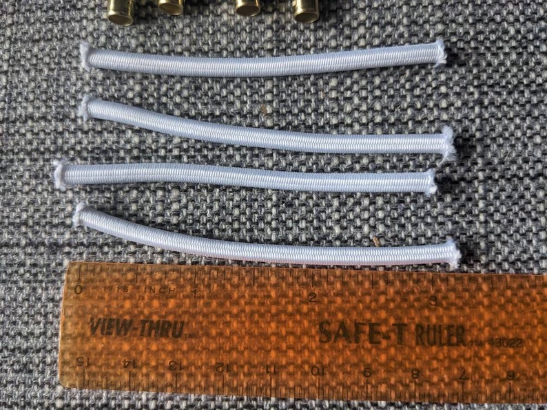 May include: Four white elastic bands laid out on a gray fabric surface with a ruler underneath. The ruler is orange and has the words "VIEW-THRU" and "SAFE-T RULER" printed on it.