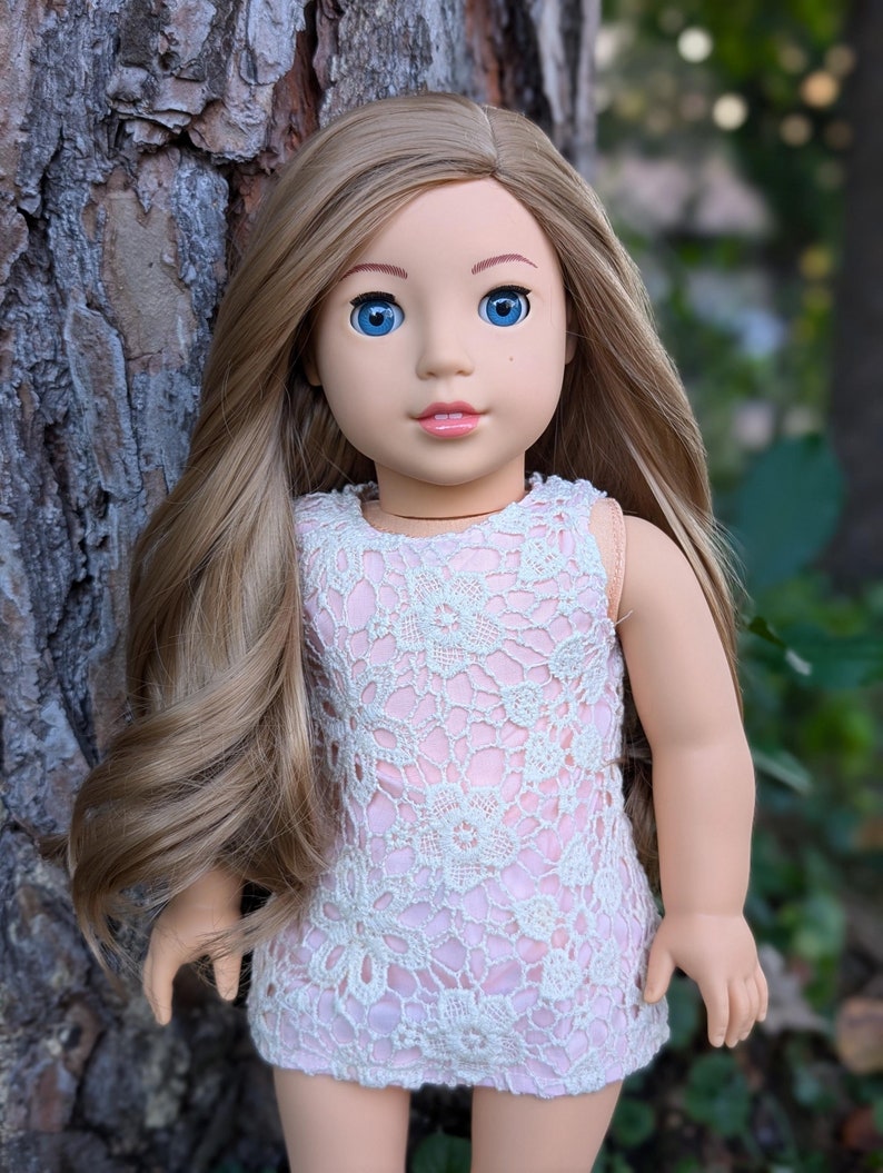 May include: A doll with long, wavy, light brown hair and blue eyes. She is wearing a pink and white lace dress.