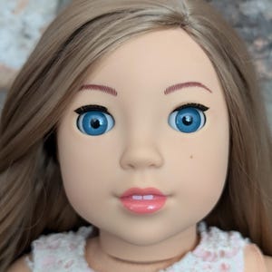 以下が含まれることがあります： 長い明るい茶色の髪と青い目をした人形。人形は白とピンクのドレスを着ています。