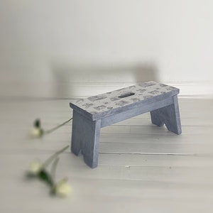 Peut inclure: Un petit tabouret en bois gris avec un dessus &agrave; motifs. Le tabouret a un design simple avec un dessus rectangulaire et quatre pieds.