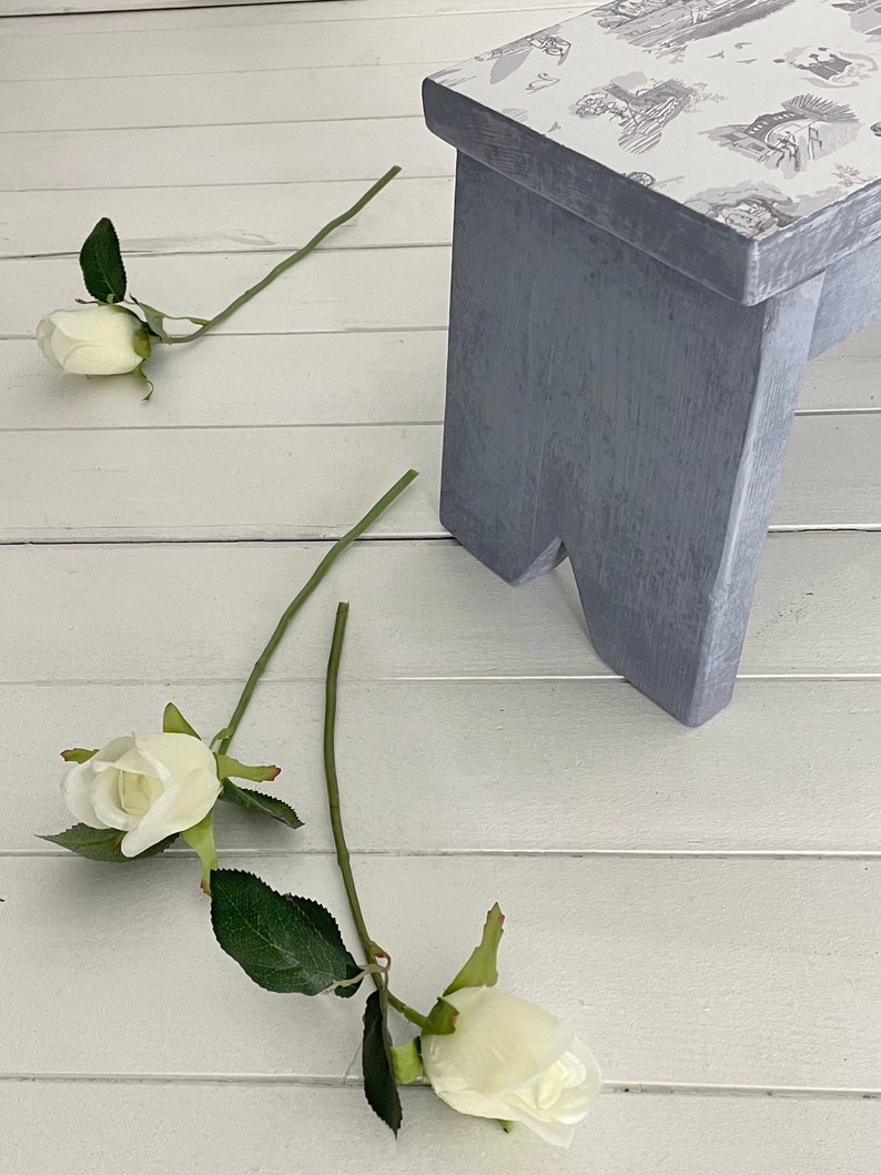 Peut inclure: Un tabouret en bois gris avec un dessus blanc orn&eacute; d'un motif floral. Trois roses blanches sont sur le sol blanc.