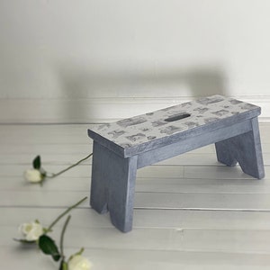 Peut inclure: Un petit tabouret en bois gris avec un dessus &agrave; motifs blancs et gris. Le tabouret est pos&eacute; sur un sol en bois blanc.
