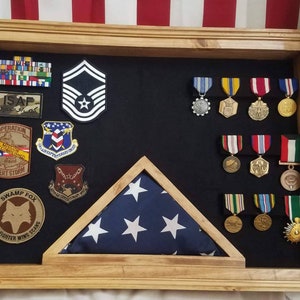 May include: A wooden shadow box display case with a folded American flag, military medals, and patches. The patches include "ISAF", "Operation Desert Storm", "Swamp Fox", and "78th Fighter Wing SCANG".