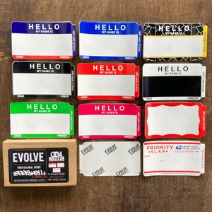 May include: A collection of name tag stickers in various colors, including blue, red, black, green, and pink. Each sticker has the text "HELLO MY NAME IS" and a blank space for writing. A box of labels is also included.