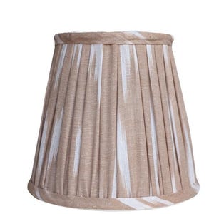 May include: A beige lampshade with vertical pleats and a white ikat pattern. The lampshade has a classic, tapered shape and is trimmed with a matching fabric. This lampshade is suitable for a table lamp or floor lamp.