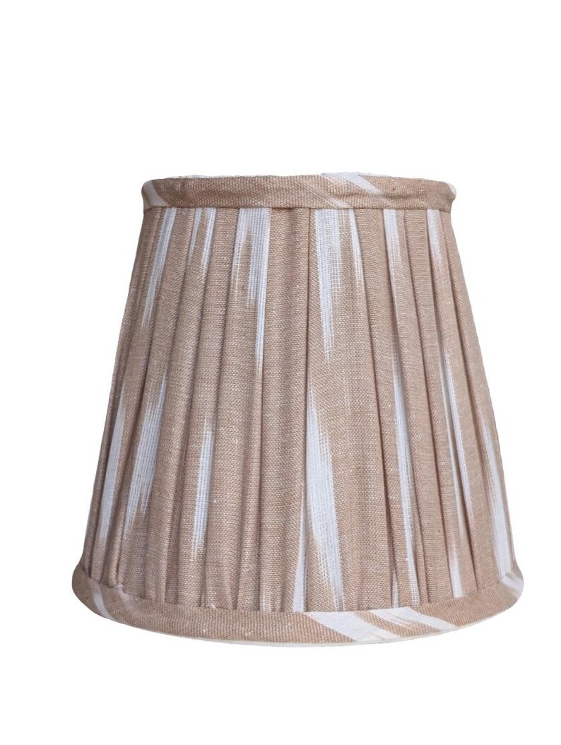 May include: A beige lampshade with vertical pleats and a white ikat pattern. The lampshade has a classic, tapered shape and is trimmed with a matching fabric. This lampshade is suitable for a table lamp or floor lamp.