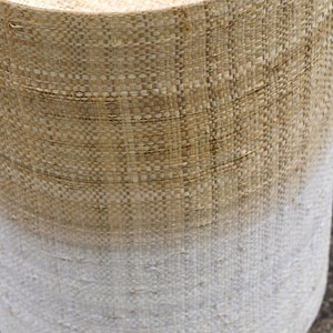 May include: A woven cylindrical stool with a natural brown top and a white bottom, creating an ombre effect.