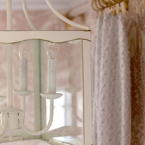 May include: White lantern chandelier with a clear glass shade and a wavy edge. The chandelier has four light bulbs and is hanging from a white ceiling.