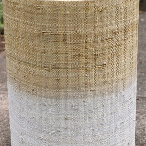 May include: A round, woven stool with a natural beige top and a white bottom. The stool is made of a woven material that gives it a rustic look.