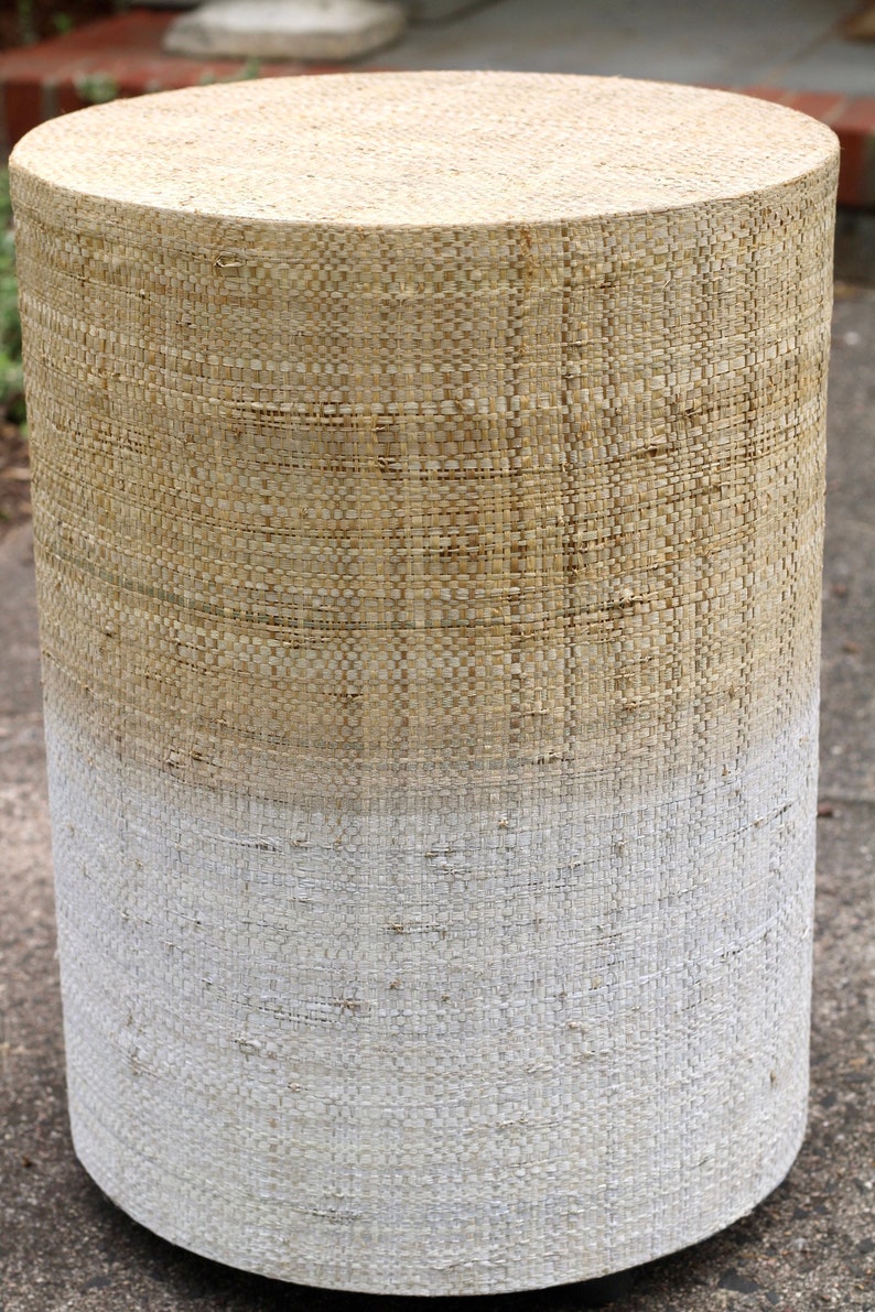 May include: A round, woven stool with a natural beige top and a white bottom. The stool is made of a woven material that gives it a rustic look.