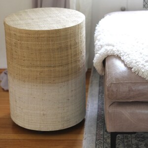 May include: A round, woven side table with a natural beige top and a white bottom. The table is sitting on a wooden floor next to a light brown leather ottoman with a white fluffy throw blanket.
