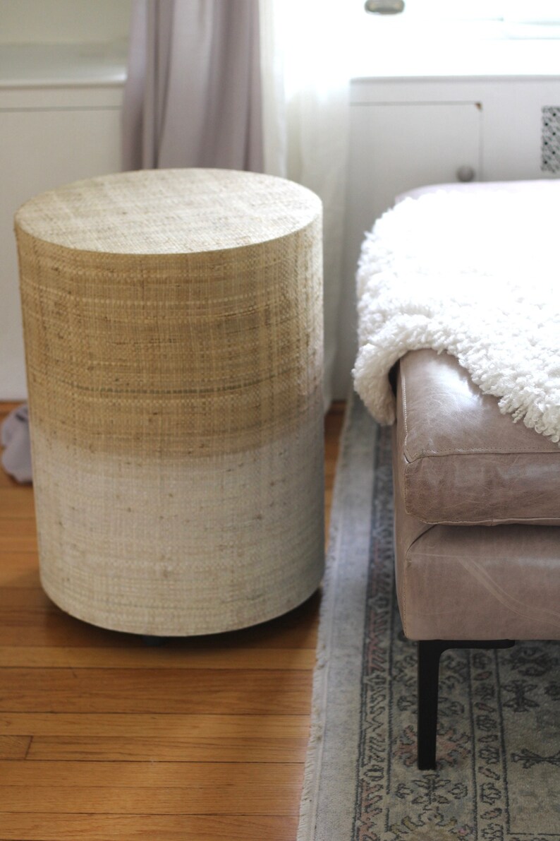 May include: A round, woven side table with a natural beige top and a white bottom. The table is sitting on a wooden floor next to a light brown leather ottoman with a white fluffy throw blanket.