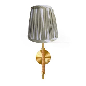 May include: A wall-mounted lamp with a pleated lampshade in shades of green and white. The lamp features a brass base and a woven rattan detail on the stem. The design is elegant and suitable for interior decor.