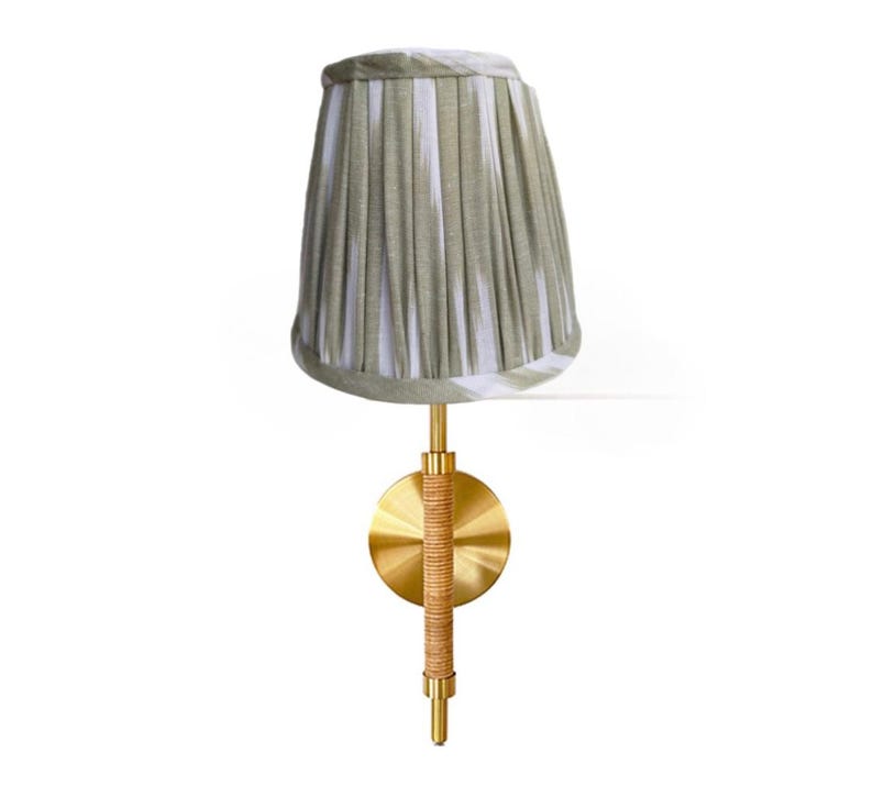 May include: A wall-mounted lamp with a pleated lampshade in shades of green and white. The lamp features a brass base and a woven rattan detail on the stem. The design is elegant and suitable for interior decor.