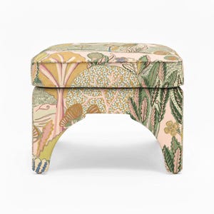 May include: A small, upholstered footstool with a botanical print. The fabric features a repeating pattern of trees, flowers, and foliage in shades of pink, green, yellow, and brown. The stool has a rectangular top and curved legs.