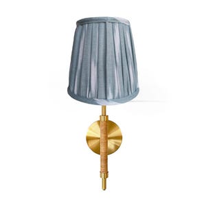 May include: A wall-mounted lamp with a pleated, light blue lampshade. The lamp features a gold-colored metal base with a circular backplate and a woven, tan-colored accent. The lamp is designed for indoor use.