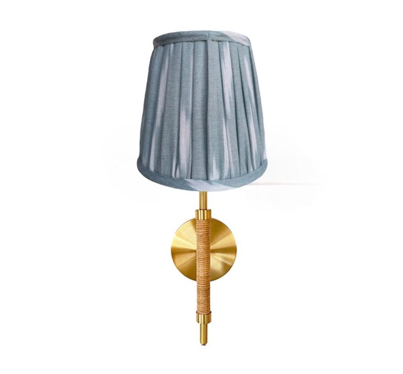 May include: A wall-mounted lamp with a pleated, light blue lampshade. The lamp features a gold-colored metal base with a circular backplate and a woven, tan-colored accent. The lamp is designed for indoor use.