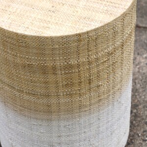 May include: A round, woven stool with a natural beige top and a white bottom. The stool is made of a woven material that gives it a rustic look.